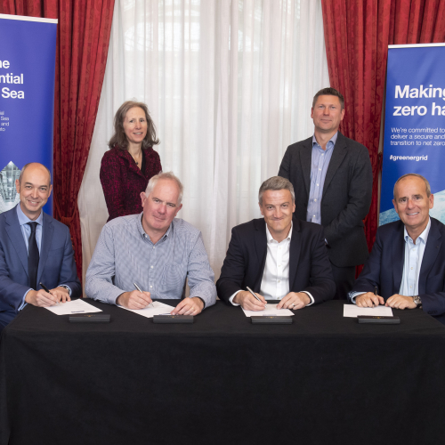 6 people stood behind a table, smiling at the camera and signing a contract