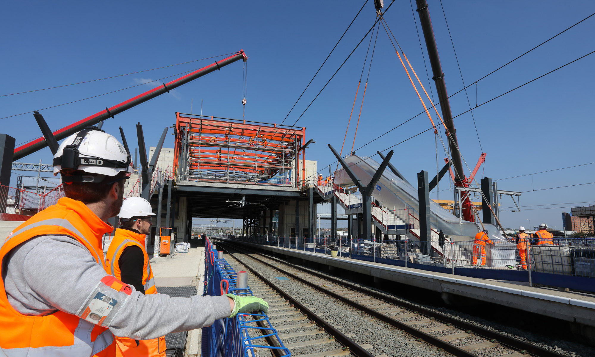 Two men in PPE watching an escalator being fitted on the other side of a rail line